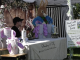 The Bloom Fest easter booth features a person in a large Easter Bunny costume standing behind a table decorated with bunny-themed buckets. A vendor sits next to the table, which has a cloth reading "Strawberry & Fig Boutique Clothing & Accessories." A decorative wooden barn backdrop and floral arrangements create a springtime atmosphere. A sign nearby advertises "Photos with the Easter Bunny" for $7 per child and an Easter egg hunt for $5, with details about times and age groups.
