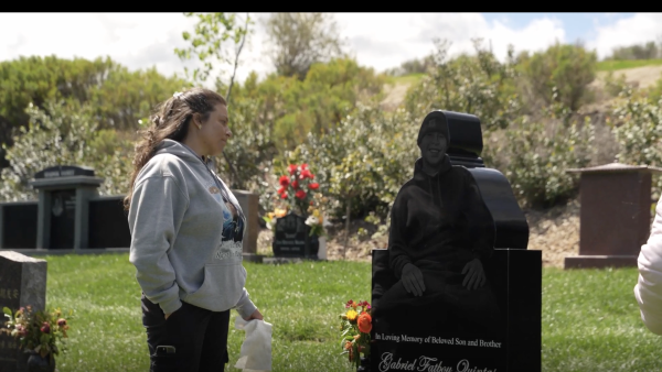 A woman stands solemnly at a gravesite in a green cemetery, holding a tissue in one hand and wearing a hoodie with a memorial image on it. She is looking at a black headstone engraved with a detailed image of a smiling man in a hoodie, along with the inscription: "In Loving Memory of Beloved Son and Brother, Gabriel Fatboy Quintas." The surrounding area features other headstones, colorful flower arrangements, and lush greenery under a partly cloudy sky.
