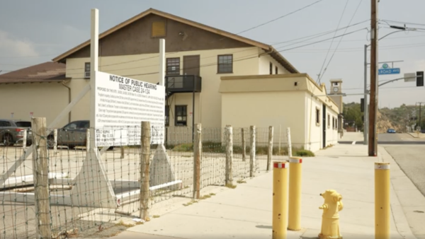 A white building with a dark-colored roof sits in the background; it is The Old Newhall Court House. The foreground features a written notice on a white board highlighting a city council meeting to discuss future development plans for the property