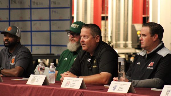 Foothill High school football coaches Sirr Guy-Shakir,Ken Holsenbeck, Dan Kelly, Jake Goossen-Brown, and sit at a table during a Foothill League Media Day panel. Name placards, water bottles, and microphones are in front of them as they address the audience.