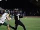 A Golden Valley High School football player in a black uniform (#5) carries the ball and stiff-arms an opposing defender in a white and green uniform (#11) during a night game on a turf field.