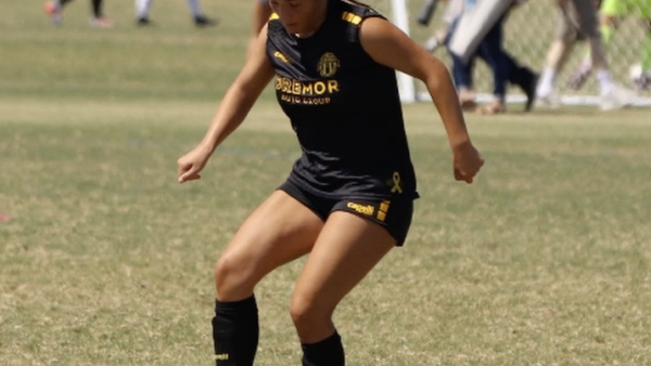 A soccer player in a black uniform with yellow lettering and accents dribbles the ball on a grass field during a match, with the goal and other players visible in the background.