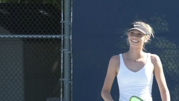 Freshman tennis player Emma Chodur’s wearing a white tank top and dark hat holding a tennis racket smiles while standing on a tennis court at college of the canyons.