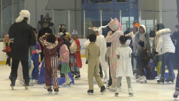 A group of children and adults in Halloween costumes ice skating at an indoor rink. Costumes include a unicorn, a witch, a ghost, a deer, a person in a judge’s robe with a white wig, and various colorful outfits. The group appears to be gathered in the middle of the rink, chatting and having fun.