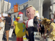 A crowd of protesters gathers outside a large government building, holding various signs and banners. Among them are people wearing costumes, including one person in a yellow chicken suit and another with a large caricature mask resembling a political figure. The scene is bright and sunny, with participants displaying messages related to political and social issues.