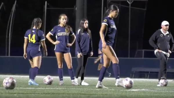 Four women on a soccer field at night prepare for practice or warm-ups. Three players wear navy uniforms with “Canyons” across the front, and one wears a navy warm-up jacket. Two soccer balls sit on the turf in front of them. One player on the right steps toward a ball, while the others stand nearby, watching or waiting. Stadium lights illuminate the scene against a dark background.
