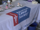 A close-up shot of a white table cloth with the American Cancer Society logo printed on a blue banner across the center. On the left side of the table, there are several small figurines of teddy bears dressed in pink and black riding silver motorcycles. The table is likely set up for a charity event.