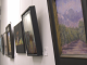 A row of framed paintings hung on a bright white wall in an art gallery or hallway. The paintings, which appear to be mostly landscapes and abstract pieces, are framed in black or dark wood and recede into the distance on the left. The painting closest to the viewer on the right depicts a green field or orchard leading up to purple mountains under a hazy sky.