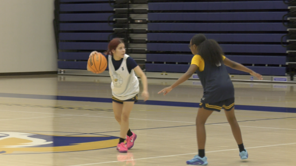 Two basketball players scrimmage on an indoor court. One player in a white practice jersey dribbles the ball while being guarded closely by another player in a dark practice jersey with yellow sleeves. The gym’s bleachers are visible in the background.