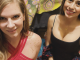 Two women smile at the camera in a warmly lit setting. The woman on the left wears a red strappy top with gold glitter on her shoulder and face. The woman on the right wears a black tank top with gold decorative designs on her face and upper chest. Colorful patterned fabric hangs in the background.