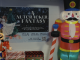 “A holiday display featuring two large nutcracker decorations—one illuminated with glittering lights on the left and a tall inflatable nutcracker on the right. Between them is a poster for ‘A Nutcracker Fantasy,’ showing a girl placing a nutcracker ornament on a Christmas tree. The poster lists showtimes for December 12 and 13 and includes a QR code for tickets. Gift-wrapped donation boxes sit below the poster, and people are visible in the background near an entrance.”