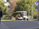 A person walks along a paved campus walkway while looking at a phone, with a maintenance golf cart driving behind them. Trees with autumn foliage, campus signage, and a blue banner line the path in the background.