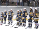 A youth girls’ hockey team stands shoulder to shoulder on the ice wearing navy and gold jerseys and white championship caps, holding their sticks during a postgame ceremony as spectators watch from the stands behind them.