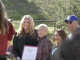 **Alt text:** A group of four people stand outdoors in a sunny, grassy area while posing for a photo. A woman in a black blazer holds a framed certificate that reads “Senate.” Beside her are an older woman with short gray hair, another woman with reddish-brown hair, and a man wearing a blue baseball cap and sunglasses. A photographer stands in the foreground taking the picture, while trees and a fence appear in the background.
