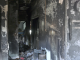 Interior hallway of a fire-damaged home with charred black walls, peeling paint, and debris scattered across the floor, with daylight visible through an opening at the far end.
