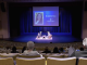 Audience members sit in a large auditorium watching a stage discussion, where two speakers are seated on chairs under blue lighting. A large screen behind them displays a portrait and the name “Dr. Jasmine Ruys” along with event branding.