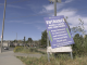 A roadside sign reading “Entrance – Haskell Canyon Bike Park Project” stands tilted on a patch of grass beside a sidewalk. The sign indicates access for contractors and authorized personnel only, with a suburban street, houses, and utility poles visible in the background under a blue sky.