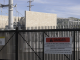 A fenced industrial facility with electrical equipment and power lines, featuring a prominent warning sign that reads “Danger: Lithium Ion Battery Storage Facility,” along with an address and emergency contact numbers. Hills and a partly cloudy sky are visible in the background.