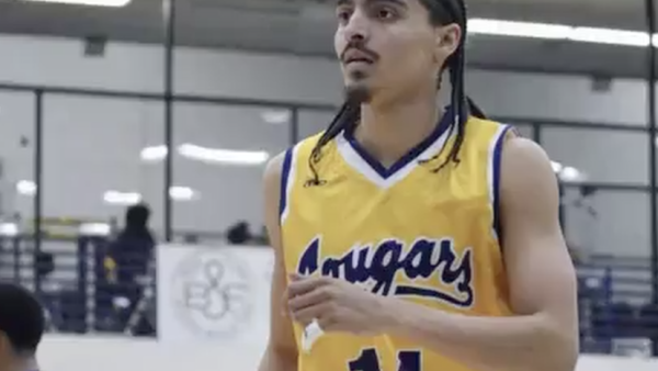 Donovan Chapman wearing a yellow “Cougars” jersey with the number 14 stands on an indoor court, looking focused during gameplay. He has braided hair and facial hair, with gym bleachers and other players visible blurred in the background.