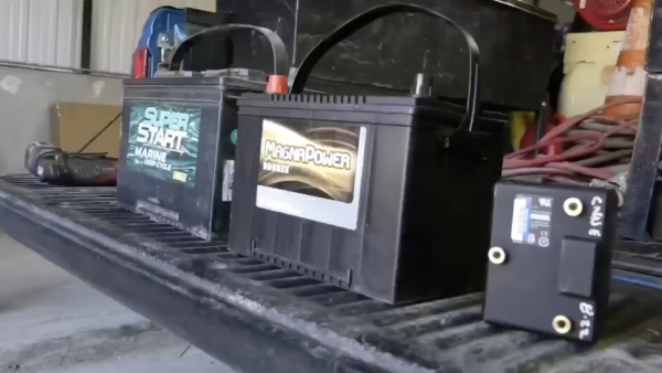 Two large black vehicle batteries, one labeled "Super Start Marine Deep Cycle" and the other "MagnaPower Bronze," sit side-by-side on the ribbed bed of a truck. To the right, a smaller black rectangular electronic component with brass terminals is also visible.
