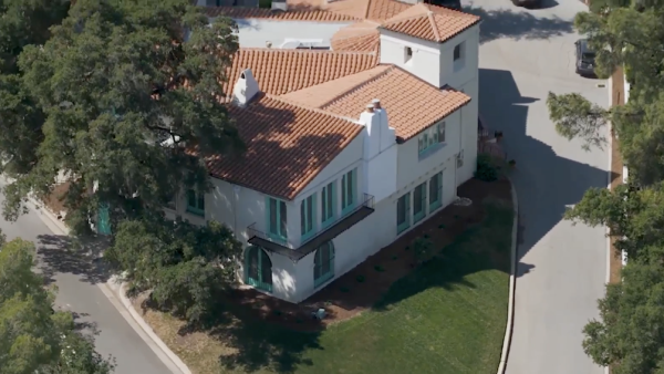 An aerial, high-angle shot of the William S Hart Museum shows a large, two-story Mediterranean-style villa with white stucco walls and a terracotta tile roof. The house features distinct teal-colored window frames and doors, including arched doorways on the ground floor and a small Juliet balcony on the second level. Two white chimneys rise from the roofline, and a square tower-like structure sits at one corner. The property is nestled between two paved roads that curve around its perimeter, surrounded by lush green trees and a small, manicured lawn.