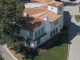 An aerial, high-angle shot of the William S Hart Museum shows a large, two-story Mediterranean-style villa with white stucco walls and a terracotta tile roof. The house features distinct teal-colored window frames and doors, including arched doorways on the ground floor and a small Juliet balcony on the second level. Two white chimneys rise from the roofline, and a square tower-like structure sits at one corner. The property is nestled between two paved roads that curve around its perimeter, surrounded by lush green trees and a small, manicured lawn.