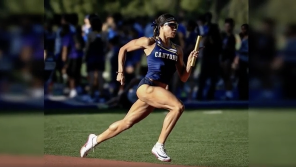 A female track athlete in a blue “Canyons” uniform sprints on a track while holding a relay baton. She is mid-stride, wearing sunglasses and a headband, with a focused expression, as a blurred crowd and other athletes stand in the background.