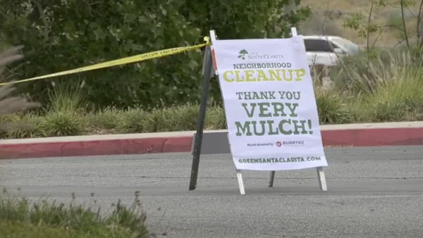 A roadside sign in Santa Clarita promotes a neighborhood cleanup event, reading “Neighborhood Cleanup — Thank You Very Mulch!” The sign stands on the edge of a street near greenery, with yellow caution tape and a parked vehicle visible in the background.
