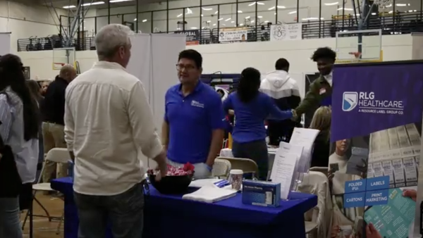 A representative wearing a blue polo shirt and glasses smiles while engaging with the visitor; next to him, a woman in a blue long-sleeved shirt is partially turned away.
