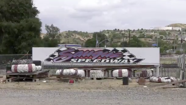 A large “Saugus Speedway” sign sits on display behind a row of worn red-and-white track barriers, with scattered debris and fencing in the foreground and rolling hills and a cloudy sky in the background.