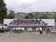 A large “Saugus Speedway” sign sits on display behind a row of worn red-and-white track barriers, with scattered debris and fencing in the foreground and rolling hills and a cloudy sky in the background.