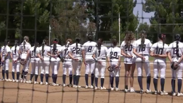 softball players in white uniforms stands shoulder to shoulder on the field during a pregame moment, seen through a protective net with trees and the outfield fence in the backgroun