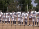 softball players in white uniforms stands shoulder to shoulder on the field during a pregame moment, seen through a protective net with trees and the outfield fence in the backgroun