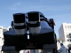 A close-up view of a telescope’s dual eyepieces points toward the sky, set up outdoors on a college campus with buildings and people gathered in the background for an astronomy event.
